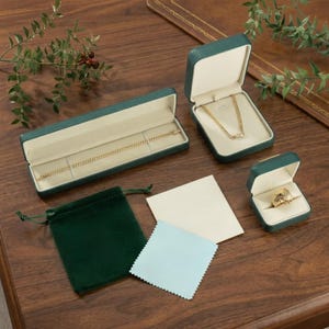 May include: An assortment of jewelry presentation items, including green velvet boxes and pouches, a blue cleaning cloth, and a gold necklace and ring. The boxes are open, displaying the jewelry against a cream-colored interior.