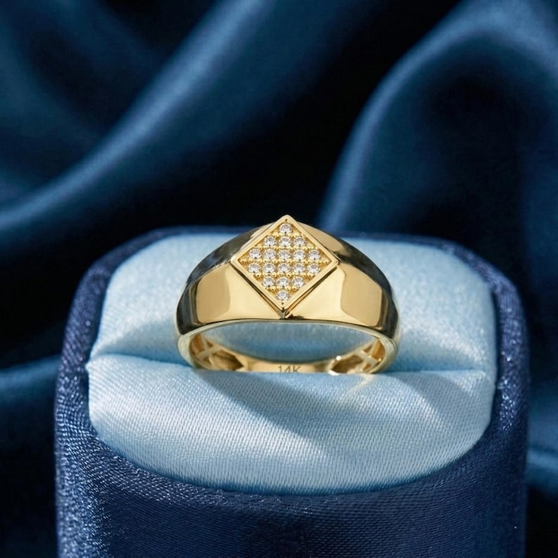May include: A gold ring featuring a diamond-shaped central design, studded with small, sparkling stones. The ring has a polished finish and is displayed against a blue backdrop, highlighting its elegant design. The ring is a piece of jewellery.