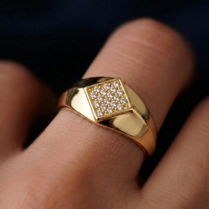 May include: A gold ring featuring a square setting adorned with small, clear gemstones. The ring has a polished surface and is displayed on a finger. The design is contemporary and stylish, appropriate for daily use or special events.