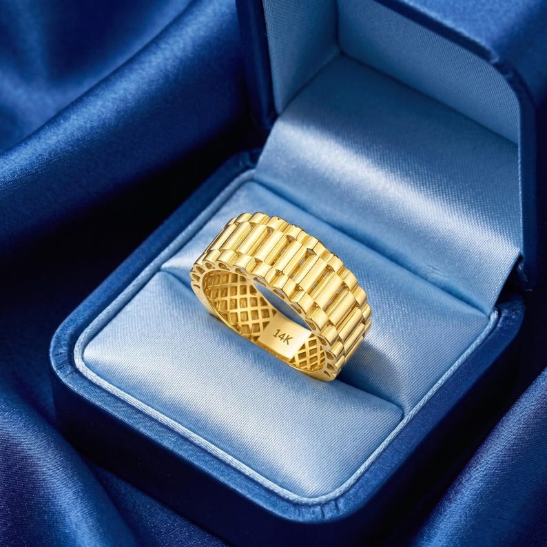 May include: A gold-colored ring with a rectangular link design, set inside a blue jewelry box. The ring is marked with "14K". The box is lined with a light blue satin material, and the background is a darker blue fabric.