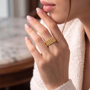 May include: A gold-colored ring with a rectangular design. The ring is worn on a finger and has a textured surface. The ring is a statement piece of jewelry.