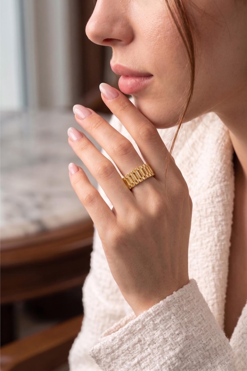 May include: A gold-colored ring with a rectangular design. The ring is worn on a finger and has a textured surface. The ring is a statement piece of jewelry.