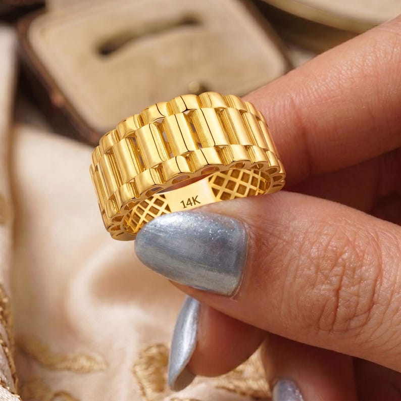 May include: A gold-colored ring with a textured, repeating rectangular design. The ring is held by a person. The ring is marked with "14K". The ring's design is reminiscent of a watch band.