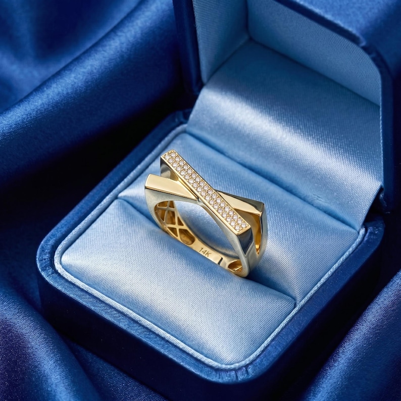 May include: A gold ring with a crisscross design, set with small diamonds. The ring is displayed in a blue velvet jewelry box. The ring is marked with "14K".