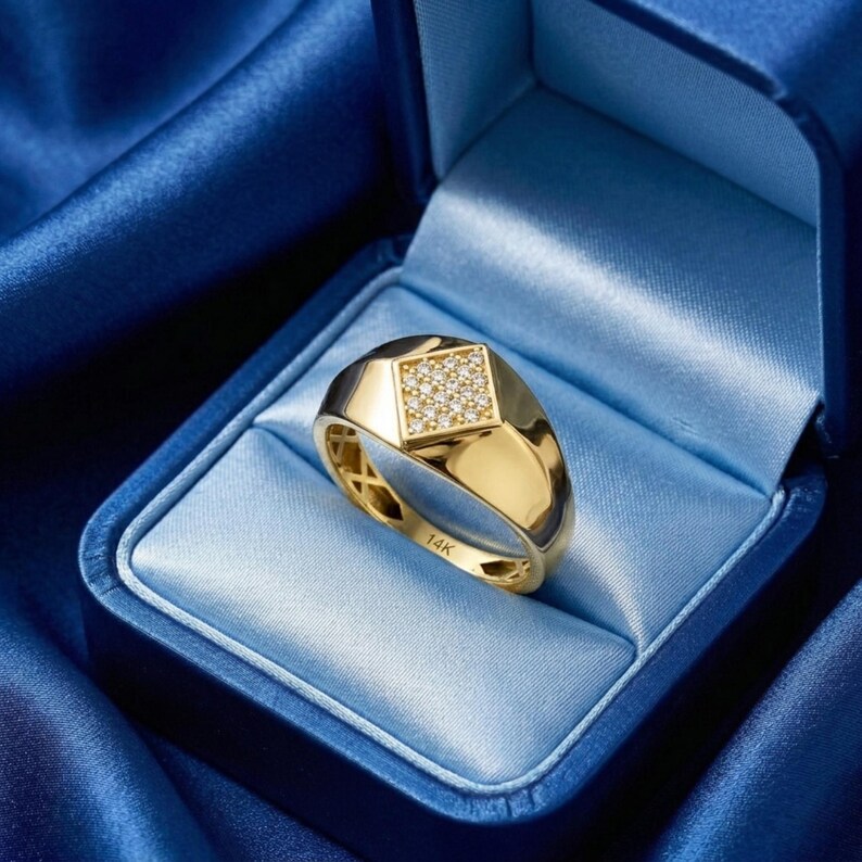 May include: A gold ring with a square diamond-studded centre, set in a blue jewellery box. The ring has a polished finish and is marked with "14K". The ring is a classic signet style.