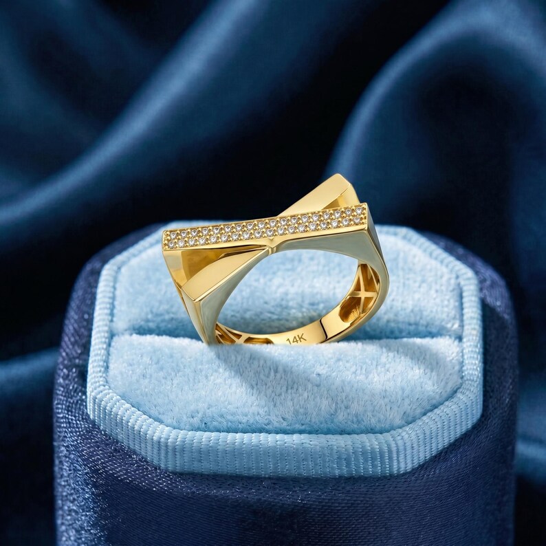 May include: A gold ring with a geometric design, featuring a row of small, clear stones. The ring is marked "14K" and sits in a blue velvet jewelry box. The ring has a modern, angular shape.