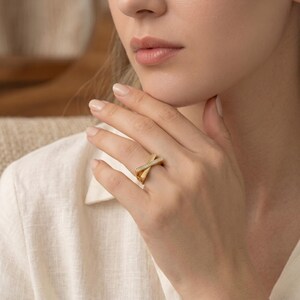 May include: A gold-colored ring with a modern, geometric design. The ring features two intersecting bands, one of which is adorned with small, sparkling stones. The ring is worn on a finger, showcasing its elegant and contemporary style.