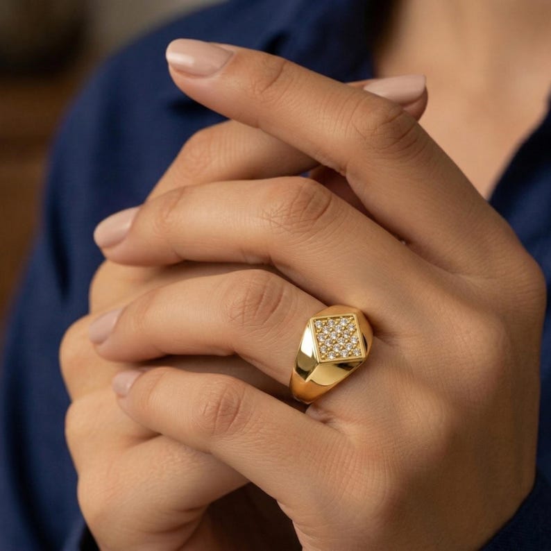 May include: A gold signet ring with a square face set with small, clear gemstones. The ring is on a finger, with the hand in front of a blue background. The ring has a polished finish.