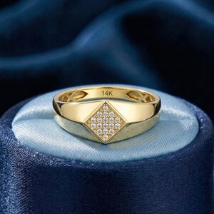 May include: A gold ring with a geometric design. The ring features a diamond-shaped centre set with small, clear gemstones. The band is a polished gold colour, and the ring is marked with "14K". The ring is displayed on a blue satin surface.