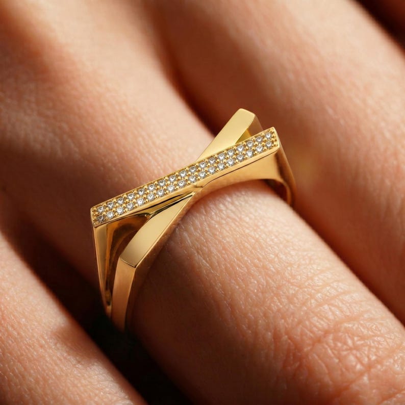 May include: A gold-colored ring with a geometric design. The ring features a band of small, clear stones set in a straight line across the top. The ring is on a finger.