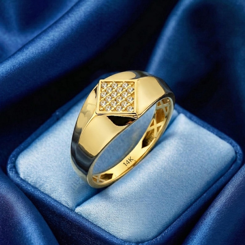 May include: A gold ring featuring a square setting of small, clear gemstones. The ring has a polished finish and is displayed against a blue backdrop. The ring is marked with "14K".