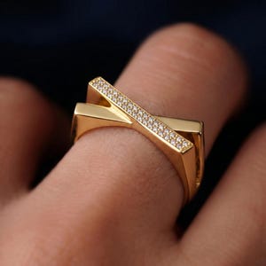May include: A gold-colored ring with a unique, geometric design. The ring features two intersecting bands, one of which is adorned with small, sparkling stones. The ring is shown on a finger, with a dark background.