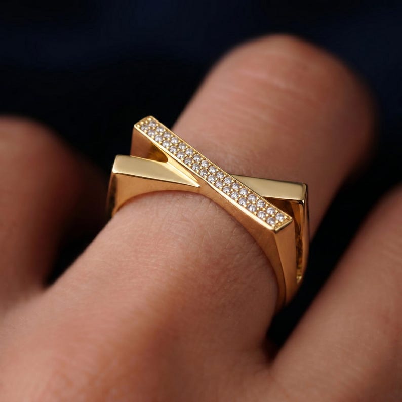 May include: A gold-colored ring with a unique, geometric design. The ring features two intersecting bands, one of which is adorned with small, sparkling stones. The ring is shown on a finger, with a dark background.