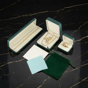 May include: An assortment of jewelry pieces, including a gold necklace, a pendant necklace, and a ring, displayed in green velvet boxes. Also included are a white and a blue cleaning cloth, and a green velvet pouch.