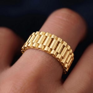 May include: A gold-colored ring with a watch band design. The ring features a series of rectangular links, creating a textured surface. The ring is worn on a finger, showcasing its bright, reflective finish.