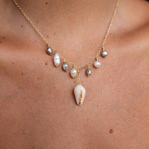 May include: A gold necklace with a fine chain, adorned with pearls. The necklace showcases a central, cream-coloured shell pendant and a collection of smaller pearls in white and grey. The necklace is displayed against a skin tone background.
