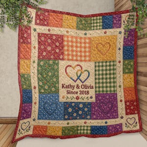 May include: A colourful patchwork quilt with squares of floral, gingham, and polka dot patterns. The centre displays two intertwined hearts with the text "Kathy & Olivia Since 2018". The quilt has a red border and embroidered heart details.