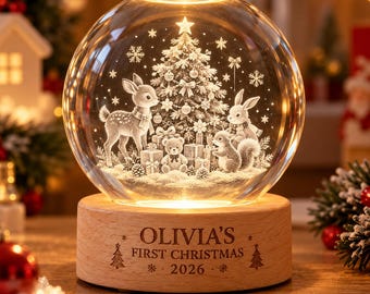 Pode incluir: Uma luz noturna em globo de vidro transparente com uma cena de Natal gravada a laser em 3D, apresentando um veado, coelho, esquilo, urso de peluche e árvore. A base de madeira redonda tem gravado "OLIVIA'S FIRST CHRISTMAS 2026".