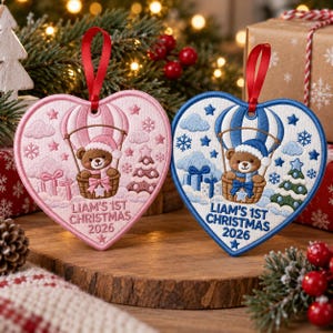 May include: Two heart-shaped embroidered ornaments with red ribbon loops. One is pink with a brown bear in a hot air balloon, the other is blue with a brown bear in a hot air balloon. Both feature the text LIAM'S 1ST CHRISTMAS 2026.