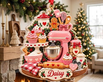 Pode incluir: Um enfeite de Natal em forma de cena de cozinha com uma batedeira rosa, cupcakes, um boneco de gengibre, um rolo de massa e um pote de biscoitos. A base inclui uma placa branca com o nome Daisy escrito em letras vermelhas.