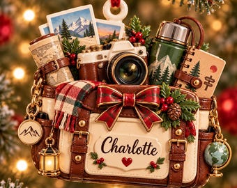 Pode incluir: Um enfeite de Natal em forma de mala de viagem castanha e creme com o nome Charlotte, uma câmara, uma garrafa térmica verde, um mapa e fotos de montanha. Decorado com um laço xadrez vermelho, azevinho e pequenos pendentes.