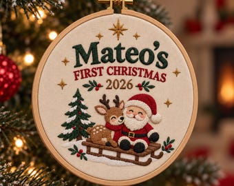 Pode incluir: Um ornamento redondo de bastidor de bordado em madeira com tecido creme bordado com o texto Mateo's FIRST CHRISTMAS 2026. O design inclui um pinheiro verde, uma rena castanha e um Pai Natal sentado num trenó de madeira.