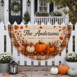 Puede incluir: Una pancarta de tela semicircular con borde a cuadros, hojas de otoño y tres calabazas. El texto dice The Andersons, Thankful, Grateful, Blessed. La pancarta está colgada en una valla blanca y tiene ojales metálicos en las esquinas.