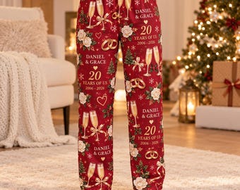 Pode incluir: Calças de pijama vermelhas com um padrão repetitivo de rosas brancas, taças de champanhe, alianças de casamento douradas e o texto Daniel & Grace, 20 Years of Us, 2006-2026. O design festivo inclui pequenos pinheiros verdes e flocos.