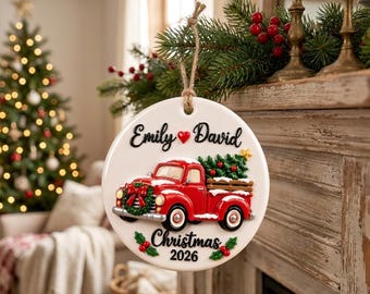 Pode incluir: Um enfeite de Natal redondo de cerâmica branca pendurado por um cordão de juta. Apresenta uma caminhonete vermelha transportando uma árvore verde, com o texto Emily e David separados por um coração vermelho e Christmas 2026 abaixo.