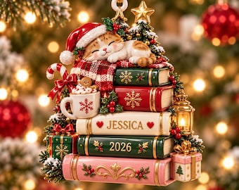 Pode incluir: Um enfeite de árvore de Natal com um gato malhado a dormir com um gorro de Pai Natal sobre uma pilha de cinco livros. Os livros têm flocos de neve, o nome JESSICA e o ano 2026. Inclui uma caneca, presentes e uma lanterna.