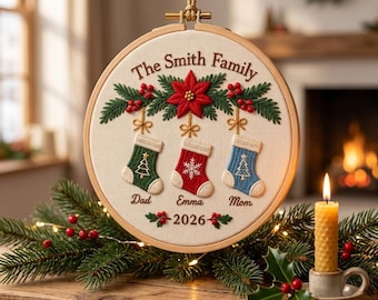 Pode incluir: Um bastidor de bordado redondo de madeira contendo tecido com decoração de Natal costurada. O texto diz The Smith Family, Dad, Emma, Mom e 2026. Apresenta uma poinsétia vermelha, ramos de pinheiro verdes, bagas vermelhas e três meias penduradas em verde, vermelho e azul com detalhes bordados em branco.