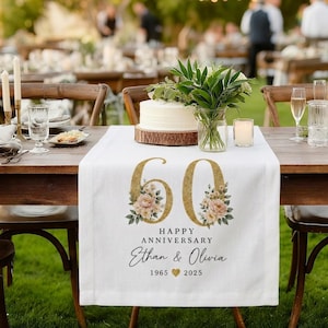Pode incluir: Um caminho de mesa de tecido branco com um número 60 grande de glitter dourado decorado com ilustrações florais. O texto diz HAPPY ANNIVERSARY, Ethan & Olivia, 1965, um ícone de coração dourado e 2025. O caminho está sobre uma mesa de madeira.