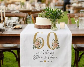 Pode incluir: Um caminho de mesa de tecido branco com um número 60 grande de glitter dourado decorado com ilustrações florais. O texto diz HAPPY ANNIVERSARY, Ethan & Olivia, 1965, um ícone de coração dourado e 2025. O caminho está sobre uma mesa de madeira.
