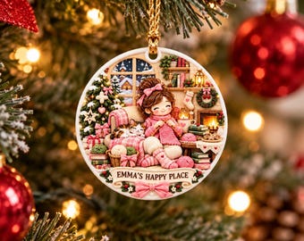 Pode incluir: Um enfeite de Natal redondo em cerâmica com uma ilustração de uma pessoa a tricotar numa sala acolhedora com um gato, lareira e árvore de Natal. O texto EMMA'S HAPPY PLACE está numa faixa na parte inferior com um laço rosa.