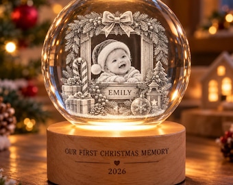 Pode incluir: Uma esfera de vidro transparente com uma imagem gravada a laser 3D de um bebé com gorro de Pai Natal, emoldurada por uma coroa, presentes e um bastão de rebuçado. O nome EMILY está gravado sob o bebé. A base de madeira tem gravado OUR FIRST CHRISTMAS MEMORY e 2026.