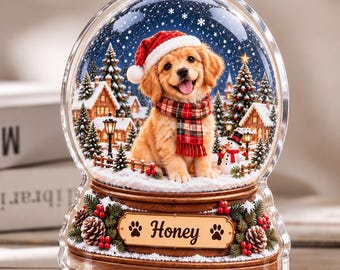 Pode incluir: Um globo de neve de acrílico transparente com um cachorrinho dourado usando um gorro de Papai Noel vermelho e um cachecol xadrez. A cena inclui uma vila nevada, pinheiros e um boneco de neve. A base marrom tem uma placa com o nome Honey.