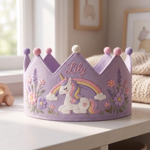 Pode incluir: Uma coroa de feltro lavanda com um unicórnio branco bordado com crina amarela e roxa, um arco-íris colorido e detalhes florais. O nome Lily está bordado em rosa na parte superior central. A coroa é decorada com pequenos pompons.