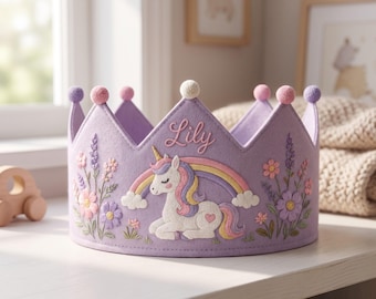 Pode incluir: Uma coroa de feltro lavanda com um unicórnio branco bordado com crina amarela e roxa, um arco-íris colorido e detalhes florais. O nome Lily está bordado em rosa na parte superior central. A coroa é decorada com pequenos pompons.