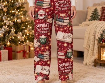 Pode incluir: Calças de pijama vermelhas com um padrão repetitivo de pilhas de livros, canecas de chocolate quente e óculos, com o texto "Sophia's Cozy Reading Season" estampado. O design inclui luzes festivas, flocos de neve e laços.