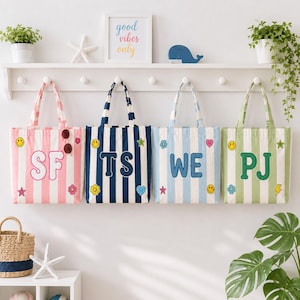 Puede incluir: Cuatro bolsas de tela colgadas en un estante de pared blanco, cada una con rayas verticales en rosa, azul marino, azul claro o verde. Cada bolsa muestra dos letras grandes, 'SF', 'TS', 'WE' o 'PJ', y está decorada con pequeños parches coloridos como estrellas y corazones.