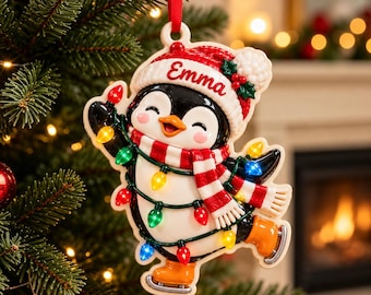 Pode incluir: Um enfeite de árvore de Natal em forma de pinguim usando um gorro de tricô vermelho e branco com o nome Emma escrito, um cachecol listrado de vermelho e branco e patins de gelo laranja, envolto em um fio de luzes natalinas coloridas.