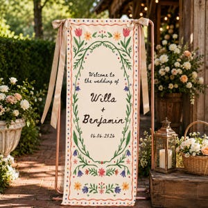 Pode incluir: Uma placa de casamento vertical em tecido creme suspensa numa estrutura de metal cor de cobre com fitas bege. A placa apresenta uma borda floral, o texto "Welcome to the wedding of Willa + Benjamin" e a data "06.06.2026" em letras escuras.