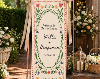 Pode incluir: Uma placa de casamento vertical em tecido creme suspensa numa estrutura de metal cor de cobre com fitas bege. A placa apresenta uma borda floral, o texto "Welcome to the wedding of Willa + Benjamin" e a data "06.06.2026" em letras escuras.