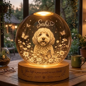 Pode incluir: Uma l&acirc;mpada memorial de vidro esf&eacute;rica com um retrato gravado de um c&atilde;o de pelo encaracolado rodeado por margaridas e borboletas. O nome Sahi est&aacute; gravado acima do c&atilde;o. A base de madeira exibe o texto You left a paw print on our hearts 2017 2026.
