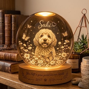 Pode incluir: Uma l&acirc;mpada esf&eacute;rica de vidro com a imagem gravada de um c&atilde;o rodeado por margaridas e borboletas. O nome Sahi est&aacute; gravado acima do c&atilde;o. A base de madeira tem a inscri&ccedil;&atilde;o You left a paw print on our hearts 2017 2026.