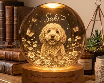 Pode incluir: Uma lâmpada esférica de vidro com a imagem gravada de um cão rodeado por margaridas e borboletas. O nome Sahi está gravado acima do cão. A base de madeira tem a inscrição You left a paw print on our hearts 2017 2026.