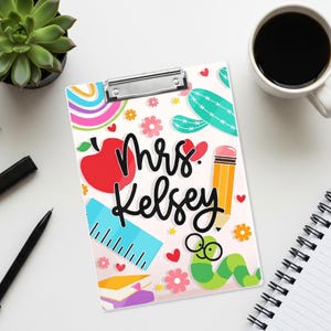 Puede incluir: Un portapapeles blanco con un clip de metal plateado que presenta un patrón colorido de una manzana roja, un lápiz amarillo, una regla azul, un gusano verde con gafas, flores, corazones y un arcoíris. El texto Mrs. Kelsey está impreso en negro.