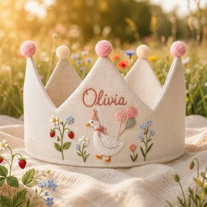 Pode incluir: Uma coroa de tecido bege com detalhes bordados, incluindo o nome Olivia, um ganso branco com um chapéu de festa, balões rosa e verdes, pequenos morangos vermelhos e várias flores azuis e brancas. A coroa é rematada com cinco pompons.