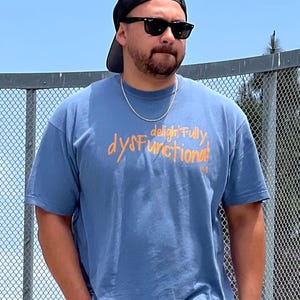May include: A blue t-shirt with the words "delightfully dysfunctional" in orange script. The person in the image is wearing a black baseball cap, sunglasses, and a silver chain necklace.