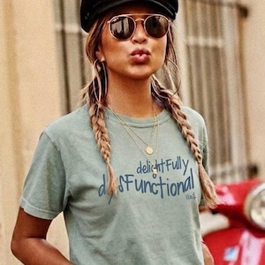 May include: A woman wearing a black cap, sunglasses, and a green t-shirt with the text "delightfully dysfunctional". She has braided hair and gold jewelry. A red scooter is partially visible in the background.